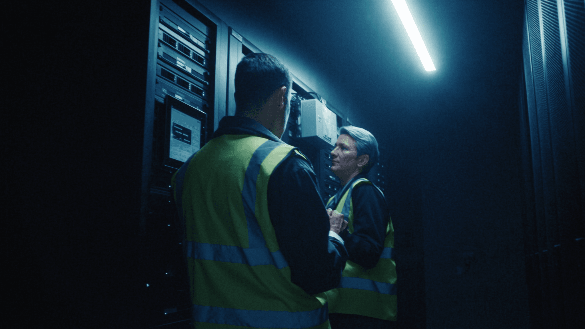 A data centre aisle with two engineers reviewing work on a tablet, sealed server cabinets on both sides lit by blue LEDs
